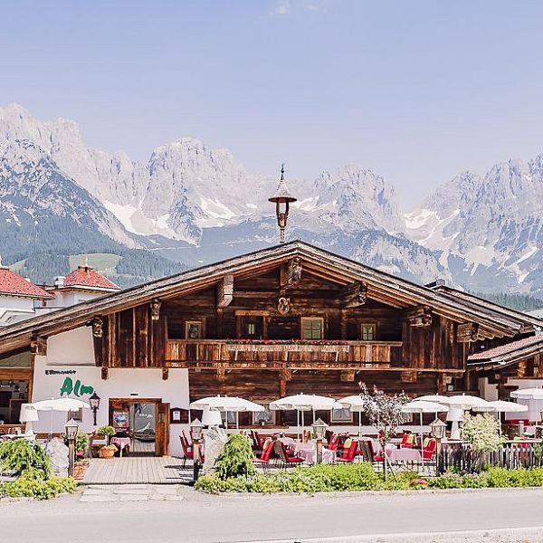 Alpenhütte mit Holzfassade und Sonnenterrasse, umgeben von malerischen Bergen. Vor der Hütte sind Tische mit weißen Sonnenschirmen aufgestellt.