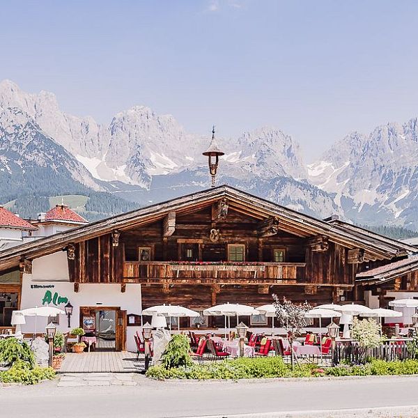 Alpenhütte mit Holzfassade und Sonnenterrasse, umgeben von malerischen Bergen. Vor der Hütte sind Tische mit weißen Sonnenschirmen aufgestellt.