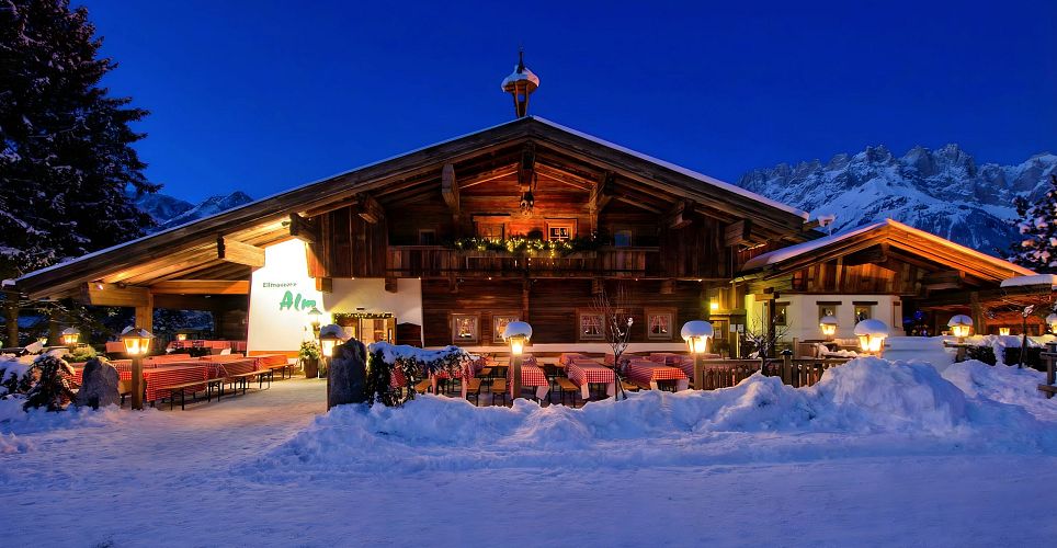 Ein rustikales Outdoor-Frühstück mit verschiedenen Speisen. Auf dem Tisch liegen Brot, Käsesorten, Aufschnitt, Marmelade und Getränke. Personen bedienen sich.