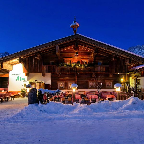 Außenaufnahme in der Dämmerung der urigen Hütte mit Tischen und viel Schnee davor