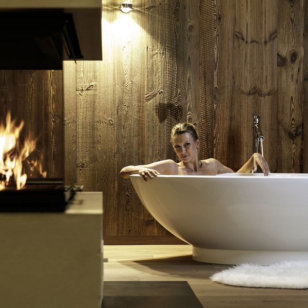 Eine Frau liegt in einer modernen Badewanne neben einem Kamin. Die Holzwand und der Teppich schaffen eine gemütliche Atmosphäre.