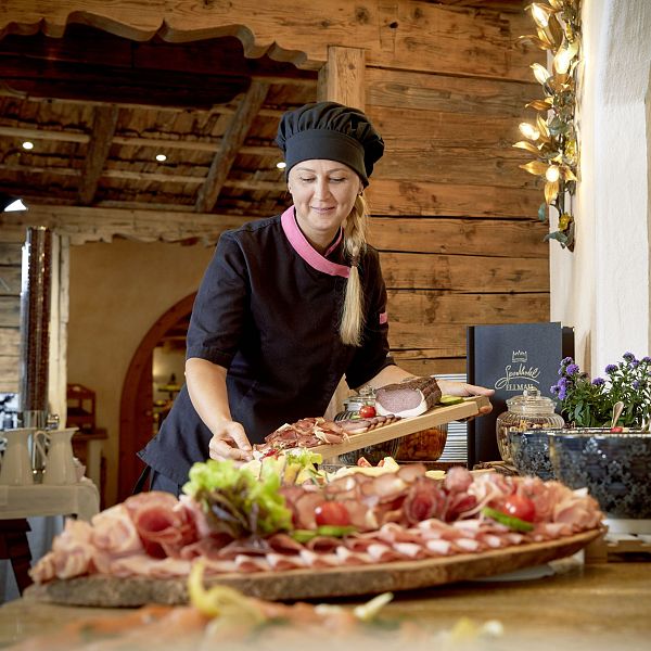 Frau in schwarzer Kochkleidung arrangiert kunstvoll Fleischplatten in einer rustikalen, gemütlichen Umgebung mit Holzwänden und warmer Beleuchtung.