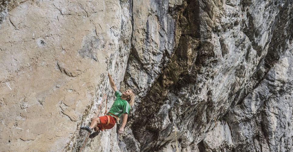 Ein Kletterer in roter Hose und grünem T-Shirt erklimmt eine beeindruckende Felswand. Das Bild zeigt den Sportler in einer herausfordernden Position.