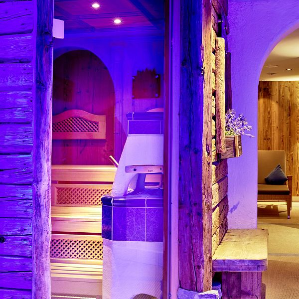 Moderner Saunaraum mit violettem Licht, Holzwänden und einer Sitzbank aus Holz. Ein Fenster mit Vorhang im Hintergrund verleiht dem Raum eine entspannte Stimmung.