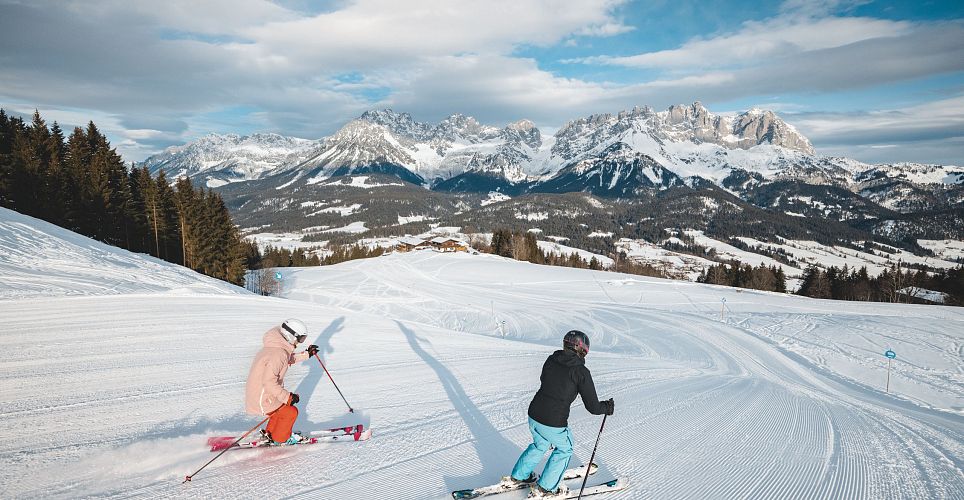 Zwei Skifahrer fahren einen schneebedeckten Hang in den malerischen Alpen herunter. Der Himmel ist bewölkt, und die umliegenden Berge bieten eine beeindruckende Kulisse.