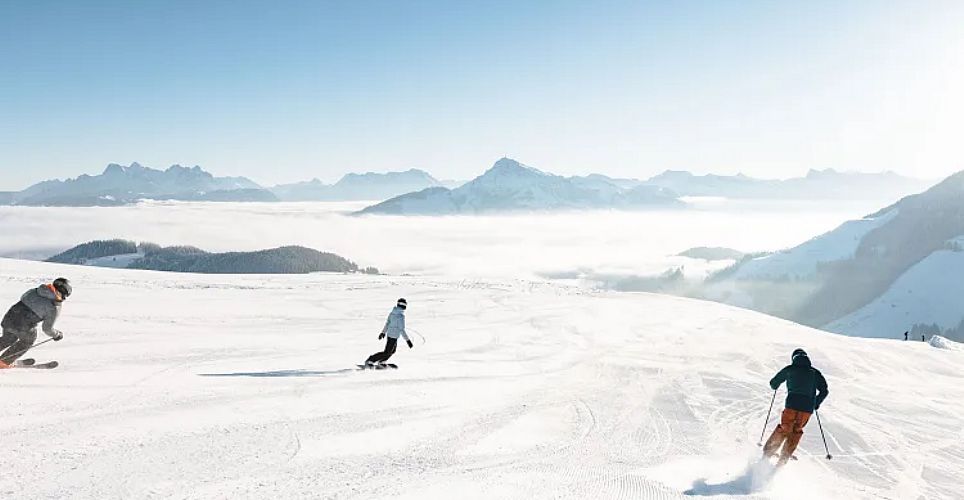 Drei Personen fahren Ski auf einer verschneiten Piste mit sonnigem Wetter. Im Hintergrund sind schneebedeckte Berge und ein klarer, blauer Himmel zu sehen.