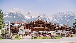 Alpenhütte mit Holzfassade und Sonnenterrasse, umgeben von malerischen Bergen. Vor der Hütte sind Tische mit weißen Sonnenschirmen aufgestellt.