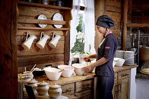 Köchin in dunkler Uniform bereitet Speisen in einer rustikalen Holzküche vor, umgeben von weißen Schüsseln und hängenden Töpfen.
