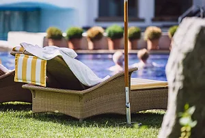 Sonnenliege mit gestreiftem Handtuch am Pool, umgeben von Pflanzen und Schwimmern im unscharfen Hintergrund. Ein ruhiges Urlaubsszenario an einem sonnigen Tag.