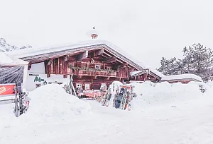 Verschneite Berghütte mit Skiern und Snowboards davor, umgeben von tiefem Schnee und Bäumen, in einem winterlichen Skigebiet.