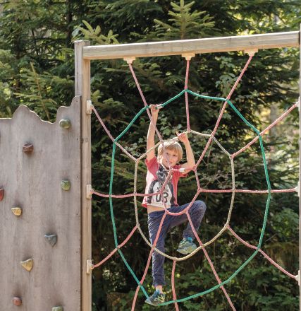 Ein Kind klettert auf einem Spielplatz an einem großen, aus Seilen geformten Spinnennetz vorbei, inmitten von Bäumen und einer Kletterwand.