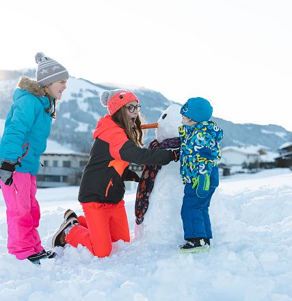 Drei Kinder bauen im Schnee einen Schneemann. Sie tragen bunte Winterkleidung, die Landschaft ist schneebedeckt mit Bergen im Hintergrund.
