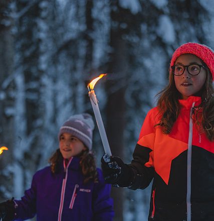Zwei Mädchen in bunter Winterkleidung halten brennende Fackeln in einem verschneiten Wald. Es ist Abend und kühl. Die Szene wirkt friedlich und winterlich.
