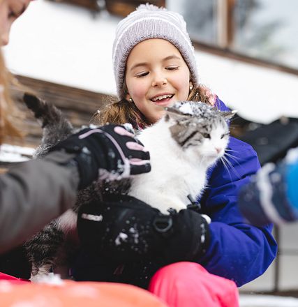 Kinder in Winterkleidung streicheln eine getigerte Katze im Schnee. Sie lachen und haben Freude beim Spiel draußen in der kühlen Winterluft.