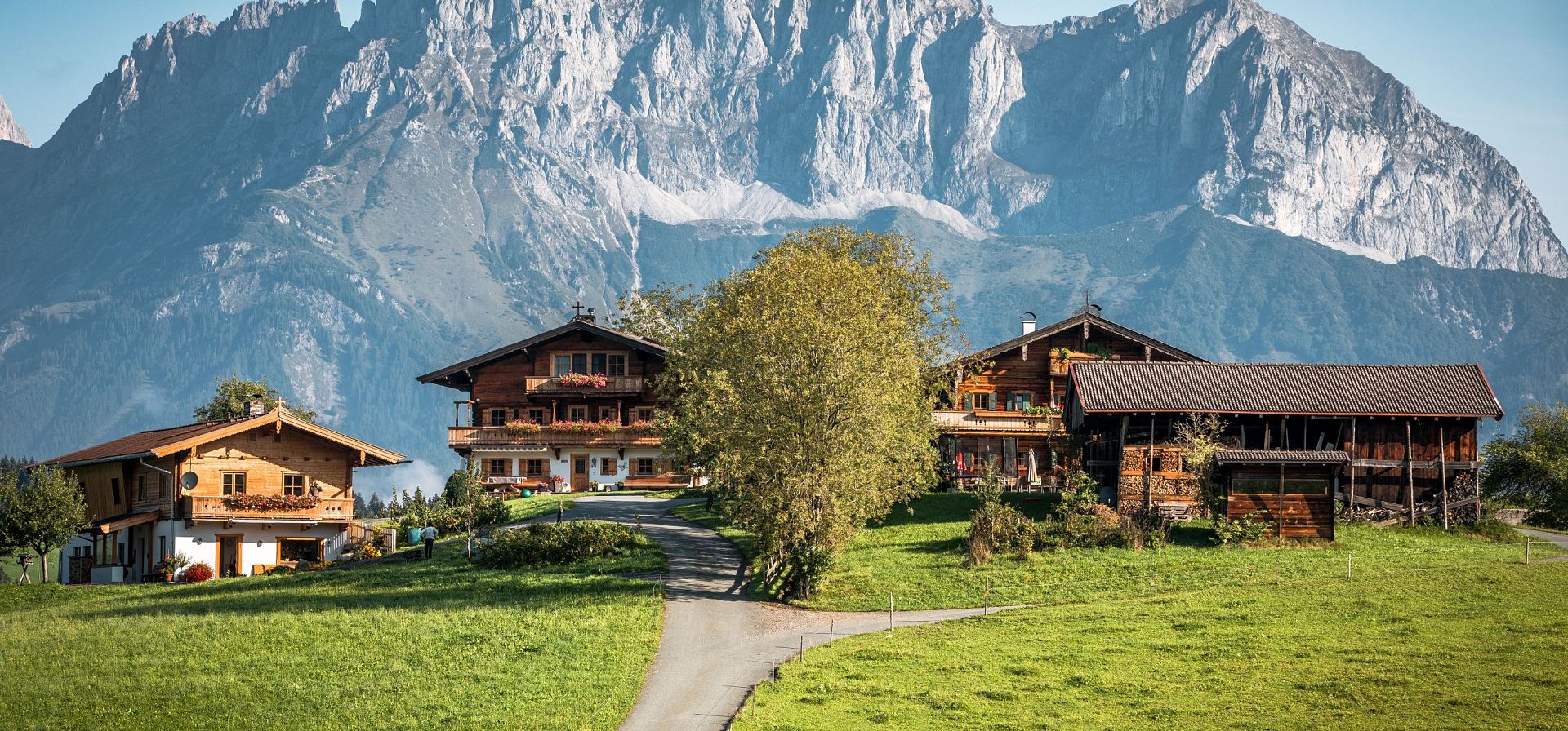 Drei Holzhäuser auf einer grünen Wiese vor dramatischen Gebirgshängen unter blauem Himmel. Ein schmaler Weg führt zu den Häusern.