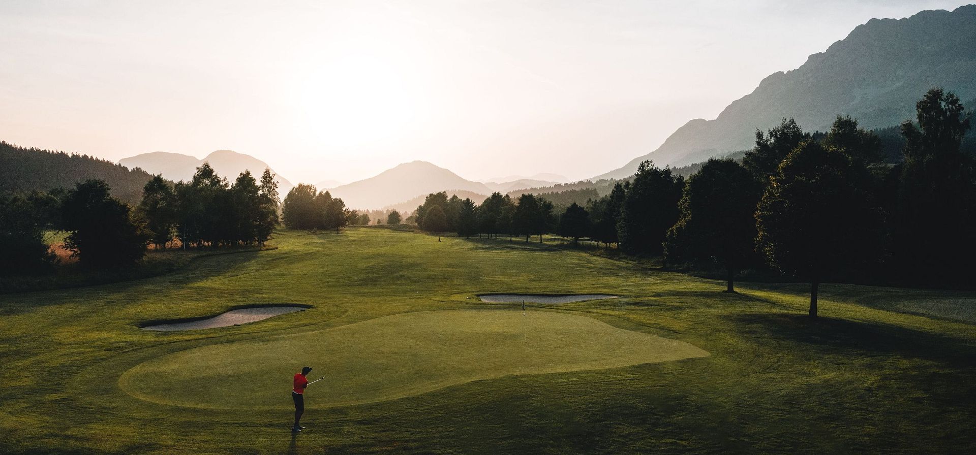 Ein Golfer steht auf einem grünen Platz, umgeben von Bäumen und Hügeln bei Sonnenuntergang, was eine ruhige und malerische Atmosphäre schafft.