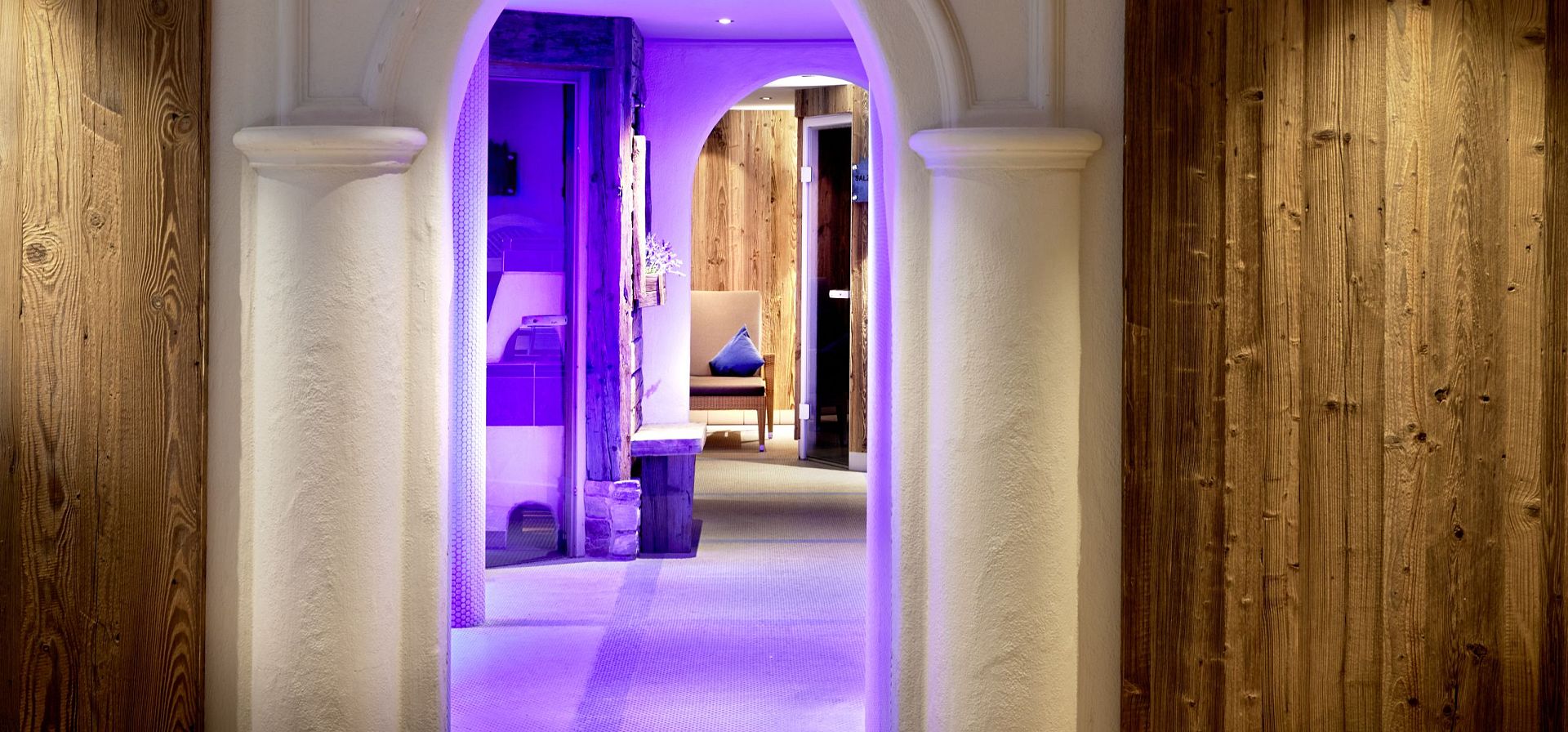 Ein stilvoll beleuchteter Eingang eines Spas mit Säulen und hölzernen Wänden, leuchtend violette Akzente schaffen eine beruhigende Atmosphäre.