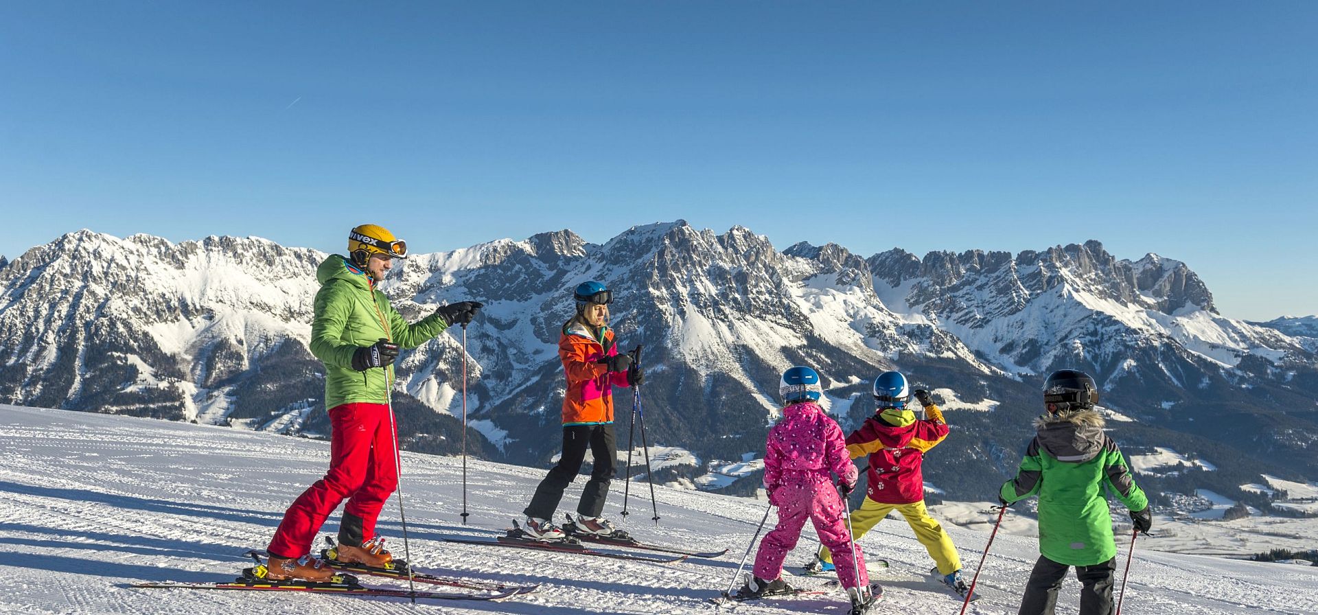 Gruppe von Kindern und Erwachsenen in bunten Skianzügen auf einer Piste in den Alpen, mit schneebedeckten Bergen im Hintergrund, unter klarem, blauem Himmel.