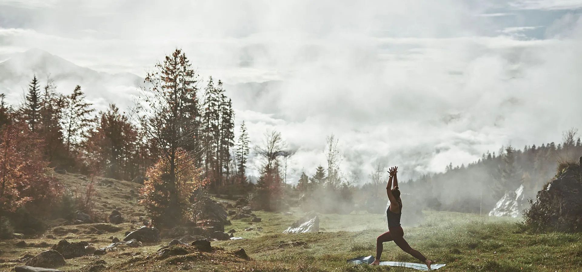 Eine Person macht Yoga auf einer Wiese in den Bergen, umgeben von Bäumen und Nebel, unter einem teilweise bewölkten Himmel.