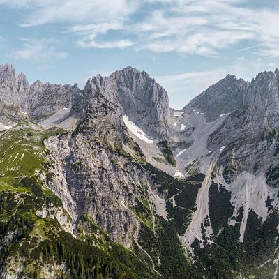 Panorama von zerklüfteten Berggipfeln mit bewaldeten und felsigen Hängen unter einem blauen, leicht bewölkten Himmel.