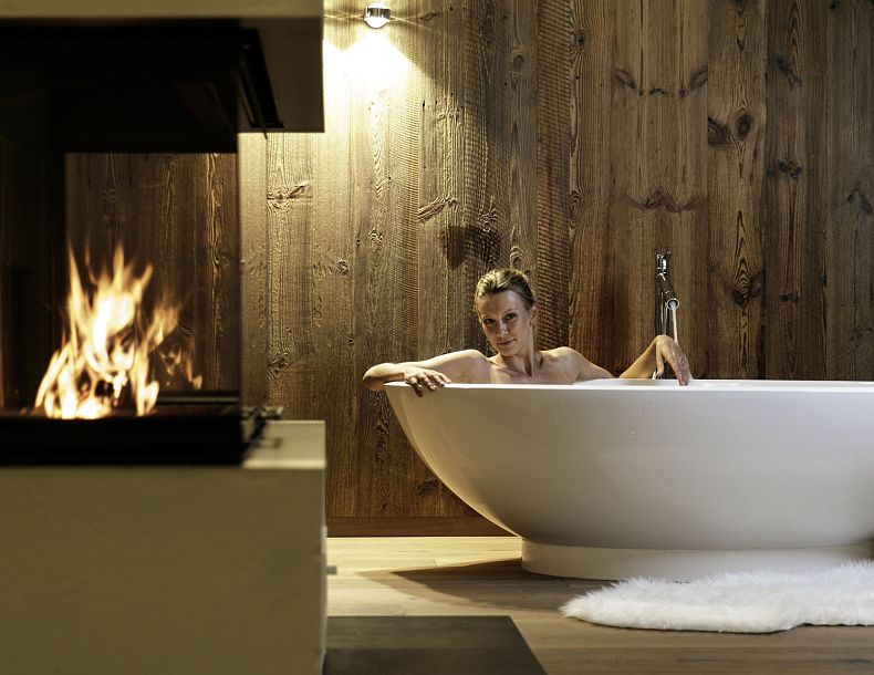 Eine Frau liegt in einer modernen Badewanne neben einem Kamin. Die Holzwand und der Teppich schaffen eine gemütliche Atmosphäre.