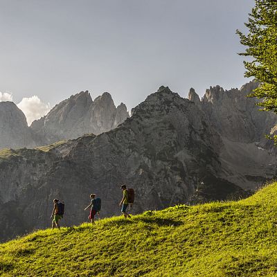 Drei Wanderer auf einem grünen Hügel vor steilen Berggipfeln unter blauem Himmel. Die Szene wirkt friedlich und majestätisch.