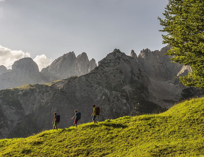 Drei Wanderer auf einem grünen Hügel vor steilen Berggipfeln unter blauem Himmel. Die Szene wirkt friedlich und majestätisch.