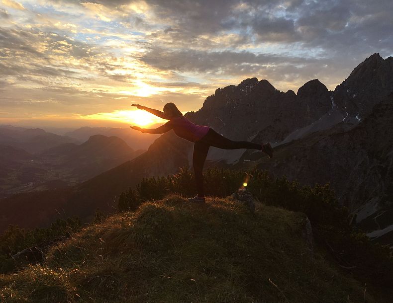 Silhouette einer Person in Yoga-Pose auf einem Hügel mit Sonnenuntergang und Bergen im Hintergrund.