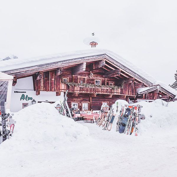 Verschneite Berghütte mit Skiern und Snowboards davor, umgeben von tiefem Schnee und Bäumen, in einem winterlichen Skigebiet.