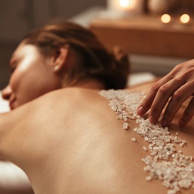 Zwei Personen entspannen auf Liegen im Spa mit Handtüchern unter den Köpfen, umgeben von Kerzen und weißen Blumen, die eine beruhigende Atmosphäre schaffen.