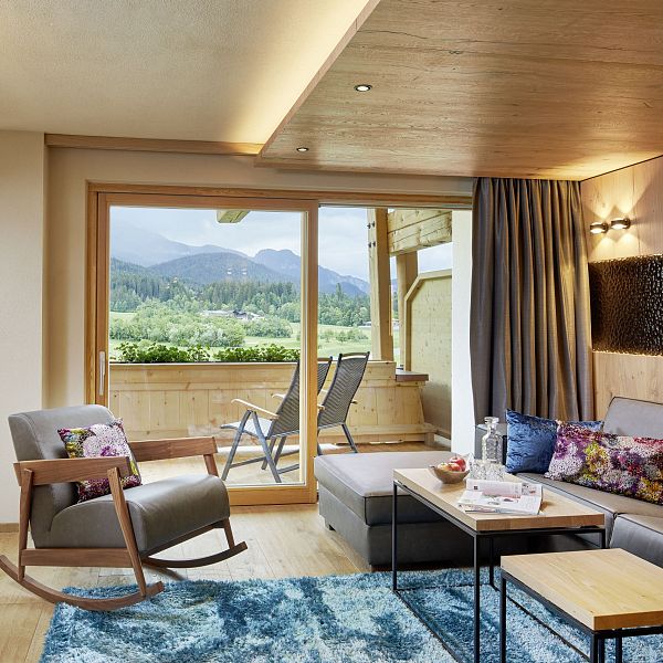 Modernes Wohnzimmer mit großen Fenstern, Holzboden und Blick auf Berge. Sofa, Schaukelstuhl und blauer Teppich gestalten den Raum gemütlich.